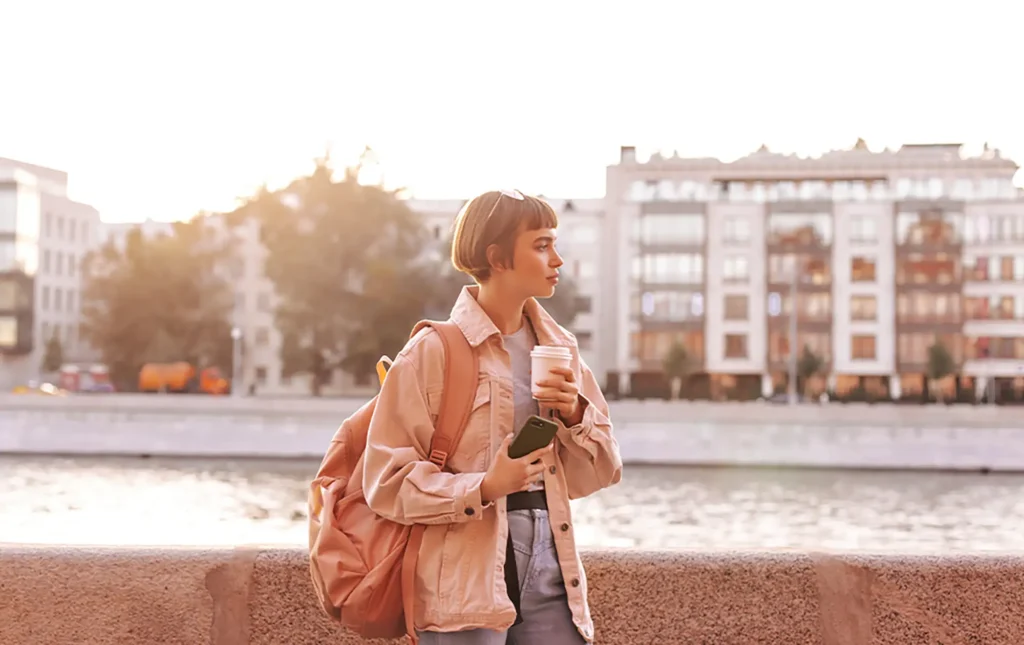 Una giovane donna con uno zaino rosa e i capelli ricci che cammina accanto a un parapetto di pietra, con edifici residenziali sullo sfondo al tramonto.