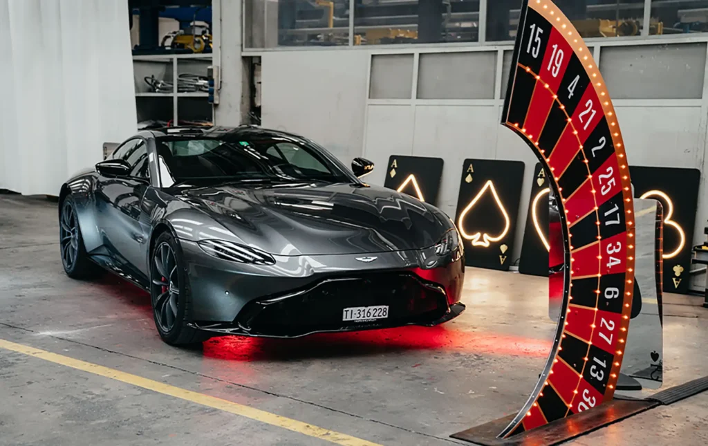 Auto sportiva nera su pedana rossa all’interno di un capannone, incorniciata da arco decorativo con simboli da casinò