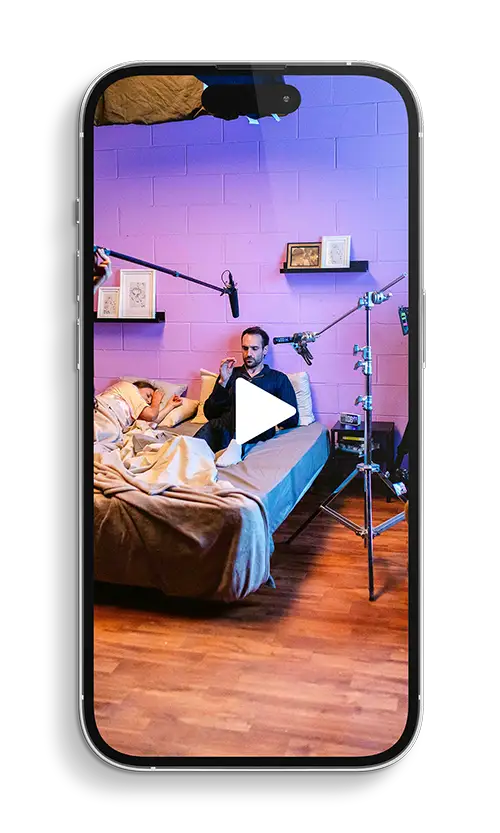 Uno smartphone moderno che mostra un video di una scena di riprese. Nel video, un uomo è seduto su un letto, mentre un'altra persona dorme accanto a lui, il tutto in una stanza con pareti viola. Sono visibili attrezzature per riprese come microfoni a boom e stativi, con un grande triangolo bianco che indica un pulsante di riproduzione al centro dello schermo.