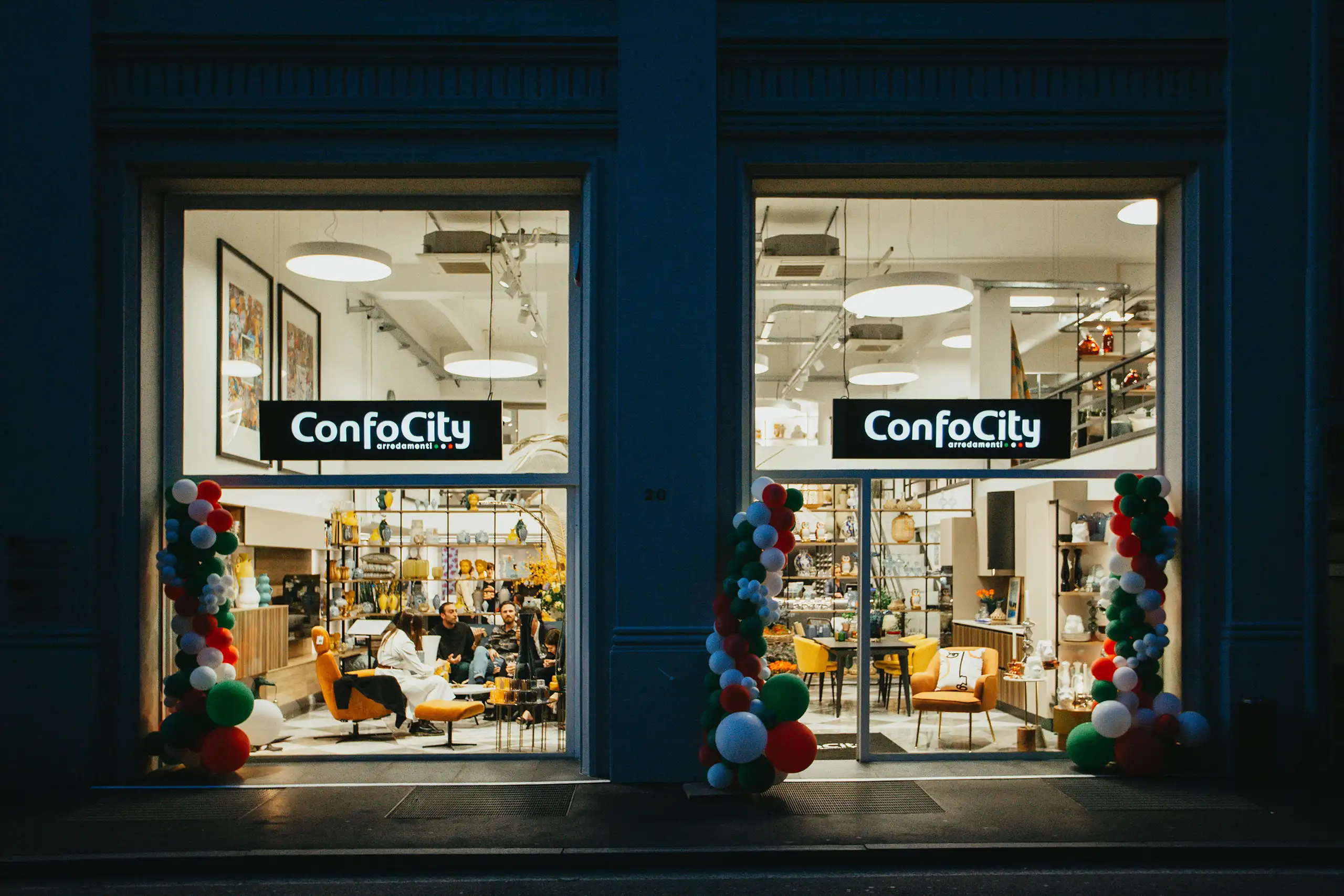 Vetrina ConfoCity di sera con logo ben visibile, arredamento in esposizione e riflessi notturni sull’ingresso illuminato.